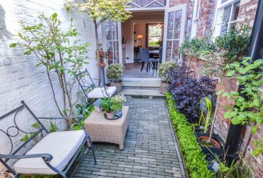 Image shows pretty, narrow rear courtyard garden with brick walls either side, ornamental trees and open patio doors showing inside the period property, for the Property Sales page of the Jameson and Partners Estate Agents website